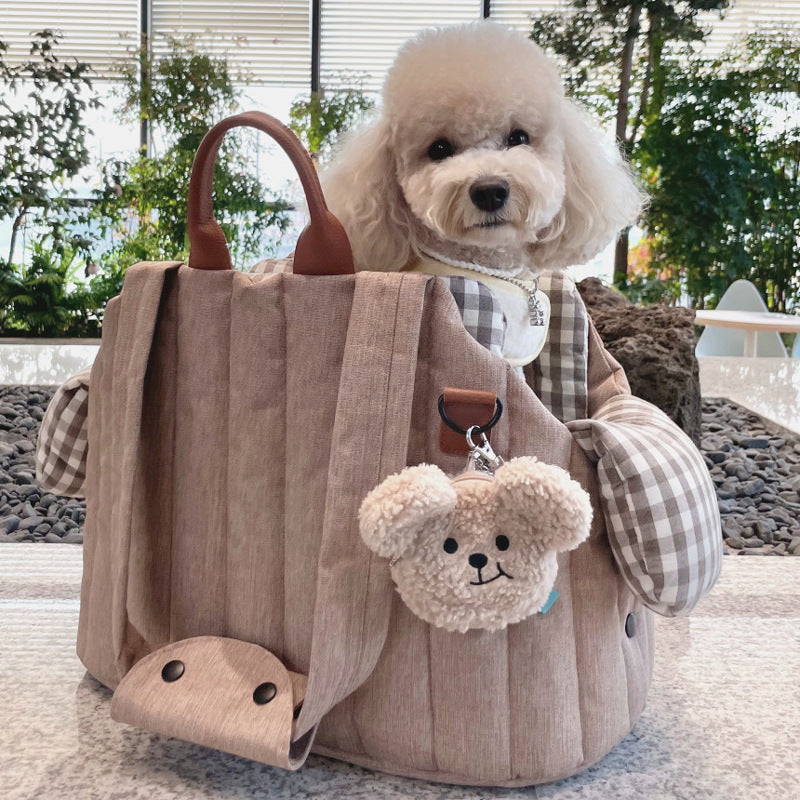 A beige pet carrier bag with a small teddy bear attached to it, designed for carrying pets. A light-colored dog is peeking out from the top of the carrier.