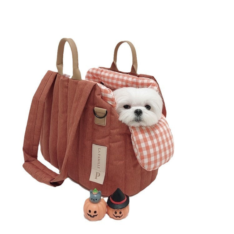 A small to medium-sized pet in a brick red pet travel bag with a checkered pattern on the flap, alongside a toy pumpkin and a bone-shaped toy.